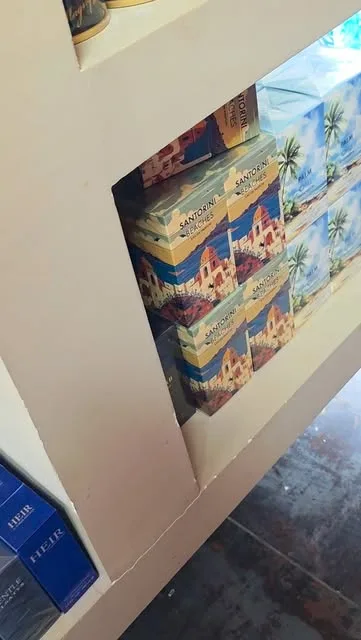 Anaquel del local visto de costado, con estuches de la edición Santorini apilados de a dos y cajas azules en la repisa de abajo.