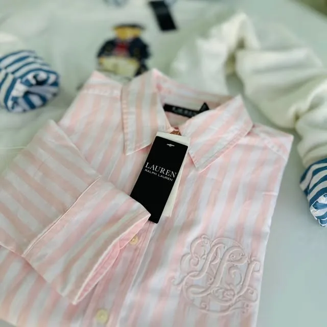 Camisa Lauren Ralph Lauren a rayas rosadas y blancas con la etiqueta puesta y un monograma bordado, sobre un suéter blanco y un oso Polo de peluche.